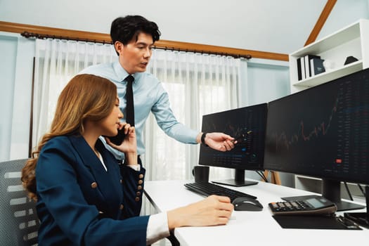 Smiling Asian businesswoman calling to broker dynamic stock exchange database with businessman partner pointing to screens comparing profit value online application program at home office. Infobahn.