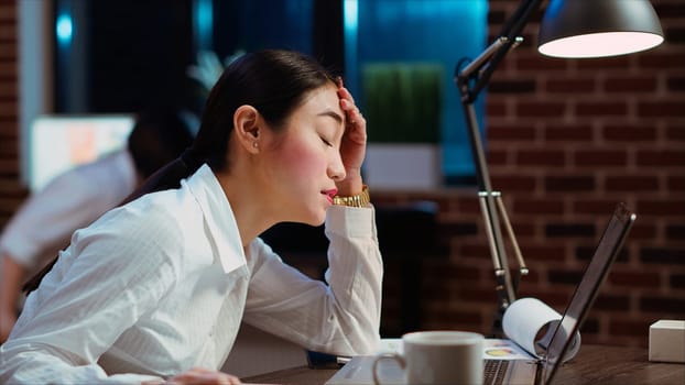 Bookkeeper feeling dread while having to do complicated tax reports in office at night. Worker squinting eyes in exasperation, feeling overworked while using laptop to do project research, camera B