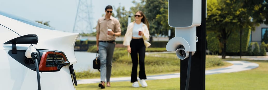 Young couple with coffee recharge EV car battery at charging station connect to electrical industrial power grid. Couple with shopping and travel using eco electric car lifestyle.Panorama Expedient