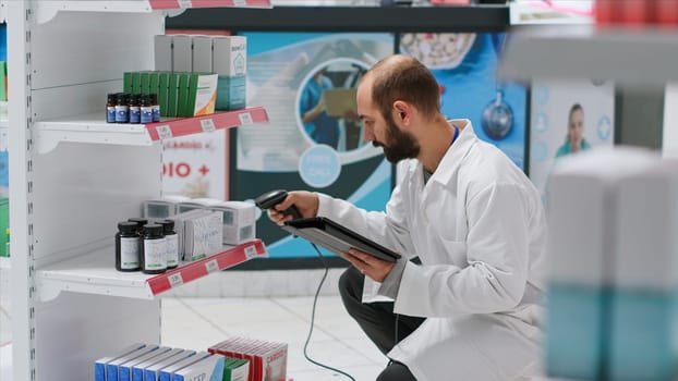 Pharmaceutical helper monitors the medication stock on shelves, scanning boxes that include prescribed medicine and cures at the clinic. Worker records medical supplies using tablet and scanner.