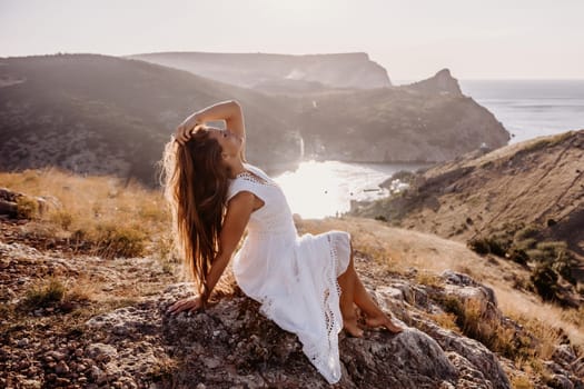 A woman in a white dress is sitting on a rock overlooking a body of water. She is enjoying the view and taking in the scenery