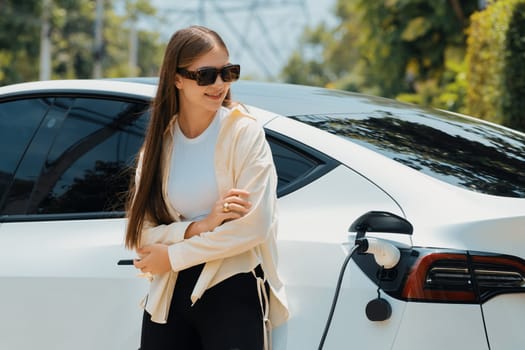 Young woman recharge her EV electric vehicle at green city park parking lot. Urban sustainability lifestyle for environmental friendly EV car with battery charging station. Expedient