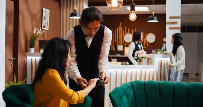 Hotel guest paying for drink and giving cash to worker, feeling happy with customer services and offering money tip. Tourist using credit card to pay at pos terminal, relaxing in lounge area.