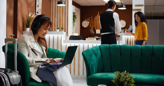 Asian woman enjoying movie on laptop in hotel lounge area, passing time waiting to be called at front desk. Young traveler using free wifi on computer, relaxing while she watches film.