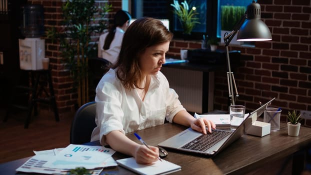 Worker writing financial business details on notepad, crosschecking with information on laptop. Businesswoman in brick wall office focused on finishing company project, camera A