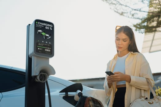 Young woman holding shopping bag and use smartphone to pay for electricity for recharging EV car battery from charging station at city mall parking lot. Modern woman go shopping by eco car. Expedient