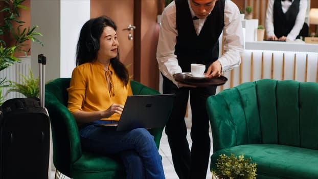 Asian woman receiving coffee while she works on laptop, sitting in lounge area at five star hotel and waiting for registration. Waiter serving drinks for guest relaxing, hospitality industry.