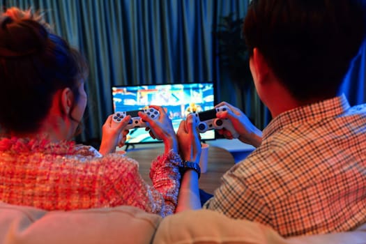 Couple gamer with controlled joystick playing fighting competition video game together on tv screen at back side view, sitting sofa at neon light colorful living room at modern comfy home. Infobahn.
