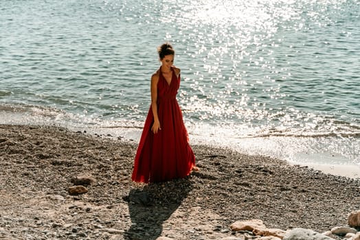 Woman red dress sea. Woman in a long red dress posing on a beach with rocks on sunny day. Girl on the nature on blue sky background