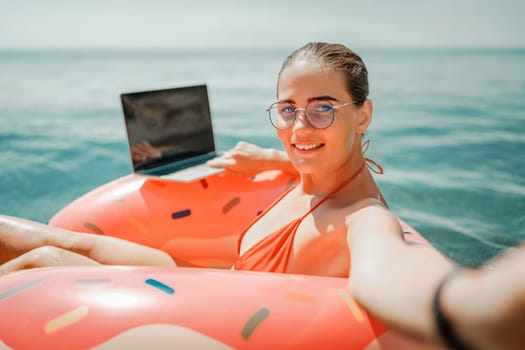 Woman laptop sea. Freelancer woman in sunglases floating on an inflatable big pink donut with a laptop in the sea. People summer vacation rest lifestyle concept