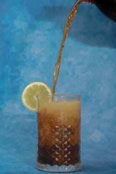 a cola is poured into a glass on a glass with ice and lemon on a blue background with copy space