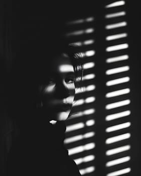 A womans face is captured in a black and white photo behind blinds, creating a pattern of tints and shades with a sense of symmetry and darkness