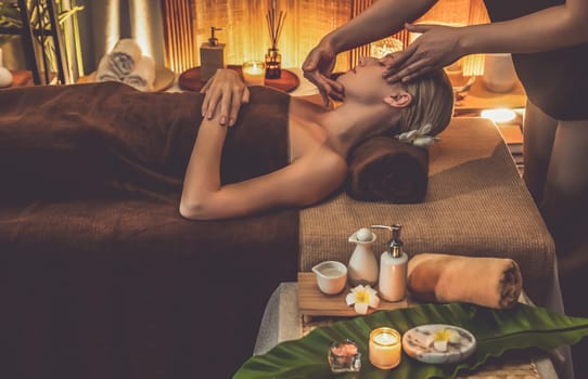 Caucasian woman enjoying relaxing anti-stress head massage and pampering facial beauty skin recreation leisure in warm candle lighting ambient salon spa in luxury resort or hotel. Quiescent