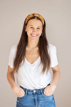 Portrait of confident beautiful woman with long brown hair, wearing casual clothes, standing in relaxed pose with hands in pockets, smiling with white teeth at camera, studio background