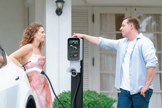 Happy and lovely couple with eco-friendly conscious recharging electric vehicle from EV home charging station. EV car technology utilized for residential home to future sustainability. Synchronos