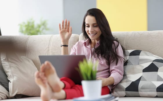 Young woman sitting on sofa and waving at laptop screen. Remote communication on social networks concept