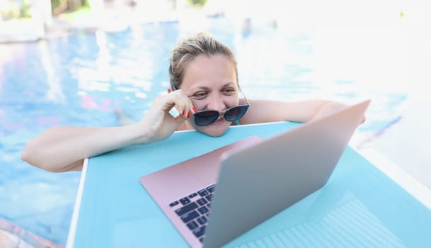 Woman straightening sunglasses and looking into laptop screen in swimming pool. Online remote communication on vacation abroad concept