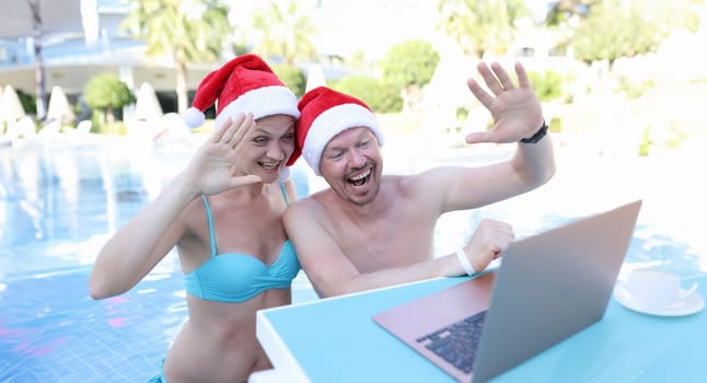 Young man and woman in santa claus hats swimming in pool and waving at laptop screen. Removed merry christmas and happy new year greetings concept