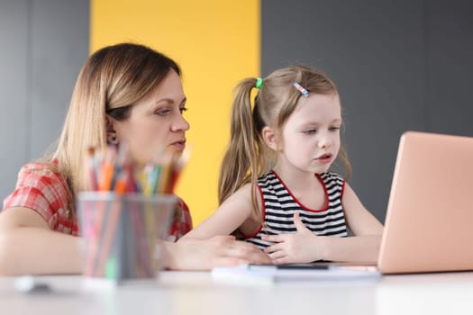 Little girl and mom are sitting at table in front of laptop screen. Online foreign language courses for children concept