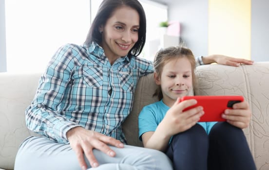 Little girl and mother are sitting on couch with mobile phone in their hands. Parental control on children gadgets concept