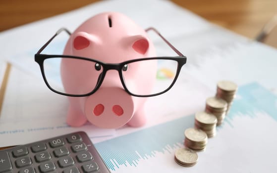 Pink piggy bank with glasses standing near heaps of coins and calculator closeup. Accounting and auditing concept