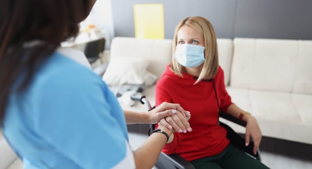 Doctor holding hand of disabled woman in wheelchair in protective medical mask. Helping people with disabilities during covid 19 pandemic concept