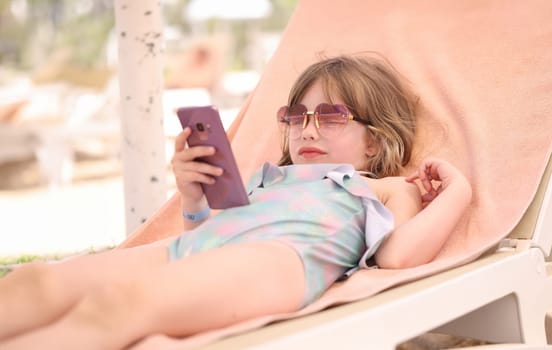 Little girl in sunglasses and swimsuit lying on lounger with mobile phone in her hands. Baby phone app concept