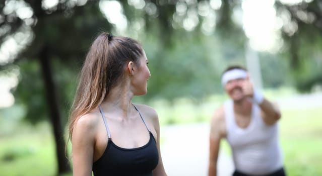 Slender girl looks back at man showing ok gesture. Adherents healthy lifestyle are running around park. People have found way to recharge their day with positive energy. Guy compliments girl.