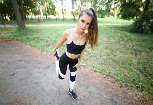 Girl in sportswear stretches leg muscles in park. After morning exercises, feeling lightness and vitality appears. Arms and legs bend slightly in joints without tension and turn them at moderate pace.