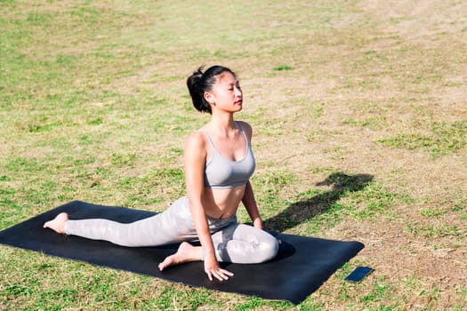 young asian woman doing stretching exercises with her yoga mat on the grass in the park using the app on her mobile phone, active and healthy lifestyle concept