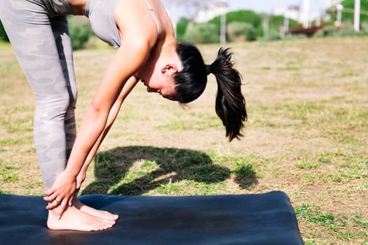 young woman doing leg stretches in the park touching her feet with her hands on a yoga mat, sport and active lifestyle concept