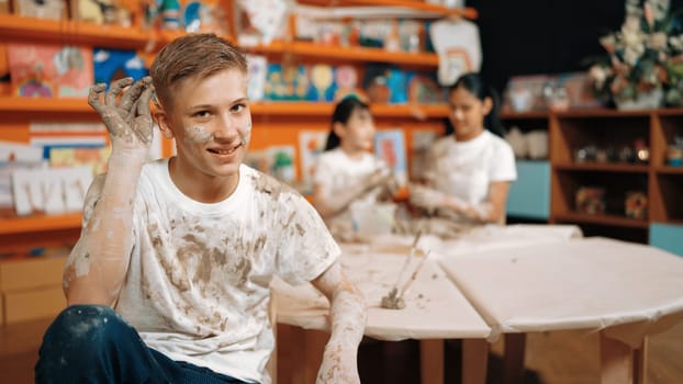 Happy student looking while put paintbrush behind ear in art lesson. Diverse student having pottery class together. Highschool boy smile while wearing white shirt with mud stained cloth. Edification.