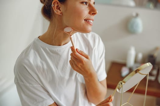 Pretty woman massaging face with jade roller and looking at round cosmetic mirror in bathroom