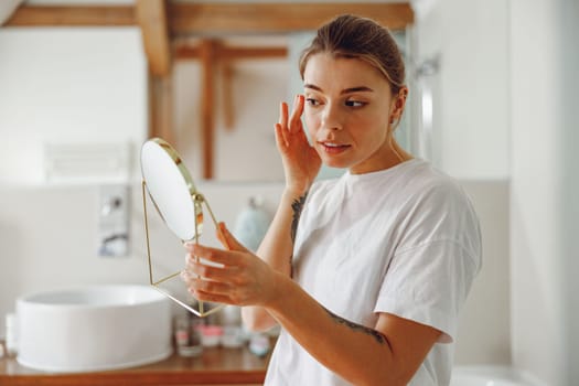 Smiling beautiful lady looking at her face in a small round mirror doing routine skincare procedures