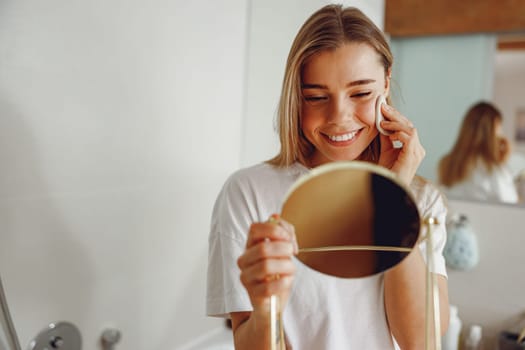 Smiling woman removing makeup with cotton pad in front of mirror standing in bathroom