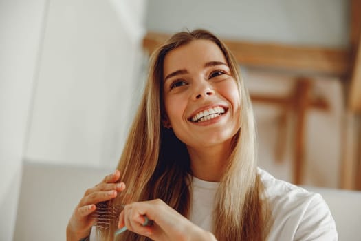 Smiling young woman brushing her hair with comb while standing in bathroom near mirror