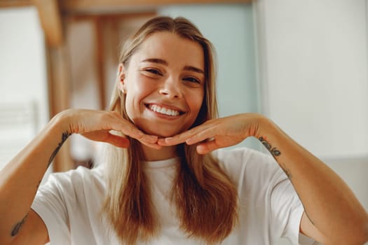 Smiling young woman touching face while standing in bathroom and looks camera