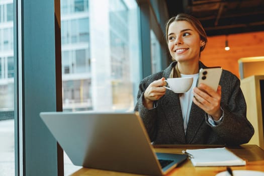 Woman manager reading message on phone and work on laptop in cafe. Distance work concept