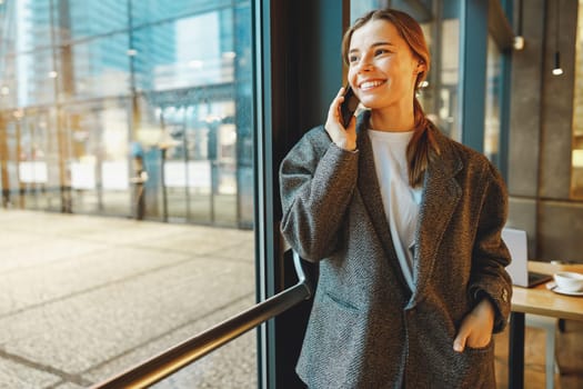 Stylish young female freelancer is talking phone with client while standing in coworking near window