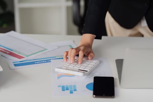 A woman is using a calculator on a desk with papers and a cell phone. The scene suggests a busy work environment where the woman is focused on her tasks