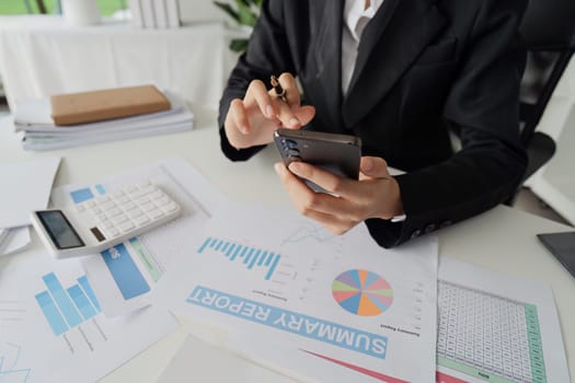 A woman in a business suit is using a calculator and a cell phone. She is focused on her work and she is in a professional setting