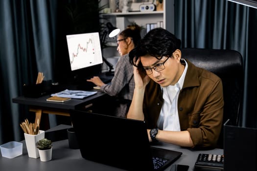 Businessman with stressful face looking on document file deadline while coworker trading stock exchange investment at late night time at modern office working on startup company project job. Infobahn.