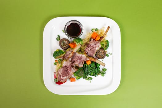 top view of a dish with juicy lamb ribs, oven-baked vegetables and fragrant red sauce on a green background