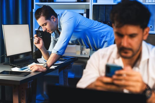 Colleagues concentrating on their job task at night home office behind desk while another man with thoughtful face solving to postpone the deadline of project at the blurred front side. Sellable.