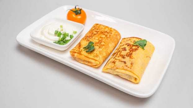 Two folded stuffed savory crepes with sour cream and chives garnish on a white plate on a white background.