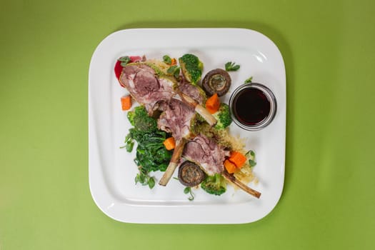 top view of a dish with juicy lamb ribs, oven-baked vegetables and fragrant red sauce on a green background