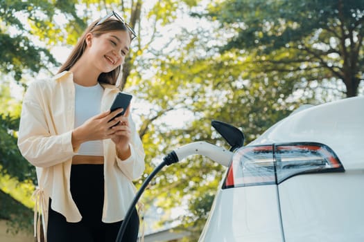 Young woman use smartphone to pay for electricity at public EV car charging station green city park. Modern environmental and sustainable urban lifestyle with EV vehicle. Expedient