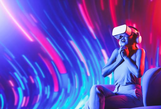 Smart female sitting on sofa surrounded by neon light wearing VR headset connecting metaverse, future cyberspace community technology. Elegant woman excited and emotionally watch movie. Hallucination.