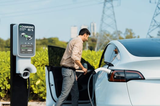 Young man recharge EV car battery at charging station connected to power grid tower electrical industrial facility as electrical industry for eco friendly vehicle utilization. Expedient