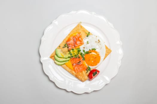 Close-up shot of a crepe with smoked salmon, avocado, egg, and tomatoes on a white plate. Square shape, elegant presentation.